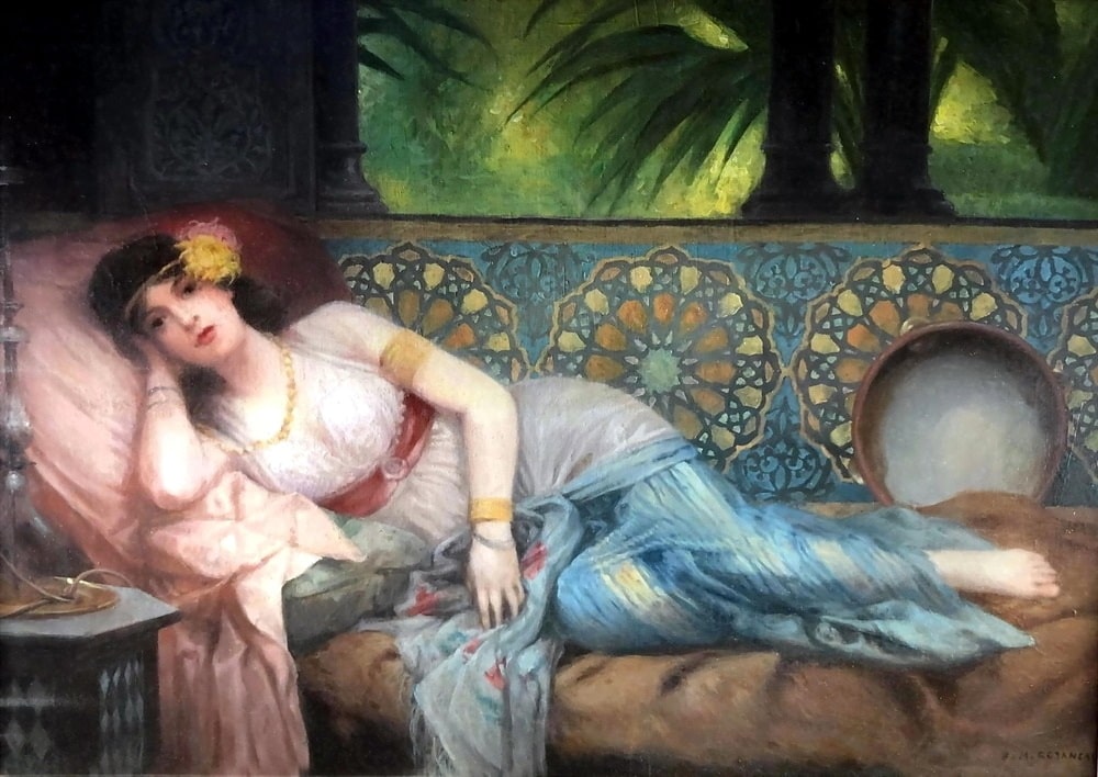 A beautiful woman with a pretty folwer in her hair is streched out on her bed with her tambourine at one end of the bed and her relaxant at the other.